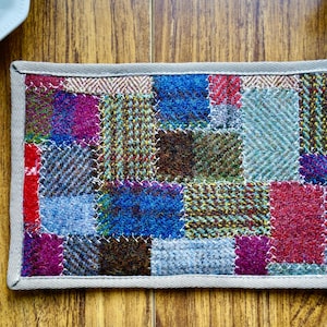 Puede incluir: Posavasos rectangular de patchwork con cuadrados de tela de tweed coloridos. El posavasos tiene un borde gris y está sobre una superficie de madera. El patchwork incluye tonos de azul, rojo, morado y verde.