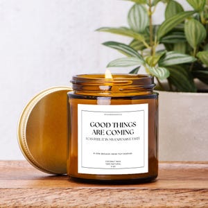 May include: A lit candle in a brown glass jar with a gold lid. The label reads "GOOD THINGS ARE COMING" and "I CAN FEEL IT IN MY EXPENSIVE TASTE". The candle is 4 oz and made of coconut wax.