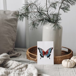 Puede incluir: Una tarjeta blanca con una ilustraci&oacute;n de mariposa azul y roja sobre un fondo blanco. La tarjeta est&aacute; sobre una bandeja de mimbre con dos calabazas blancas.