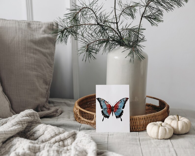 Puede incluir: Una tarjeta blanca con una ilustraci&oacute;n de mariposa azul y roja sobre un fondo blanco. La tarjeta est&aacute; sobre una bandeja de mimbre con dos calabazas blancas.