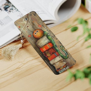 May include: A colorful bookmark featuring a cartoon caterpillar with large eyes, perched on top of a stack of books. The caterpillar is green with a brown head and has a whimsical, cartoonish style.