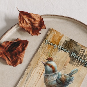 Puede incluir: Una tarjeta de estilo vintage con una ilustraci&oacute;n de un p&aacute;jaro azul y verde y el texto "Sing Your Melody". La tarjeta est&aacute; sobre un plato beige con dos hojas marrones.
