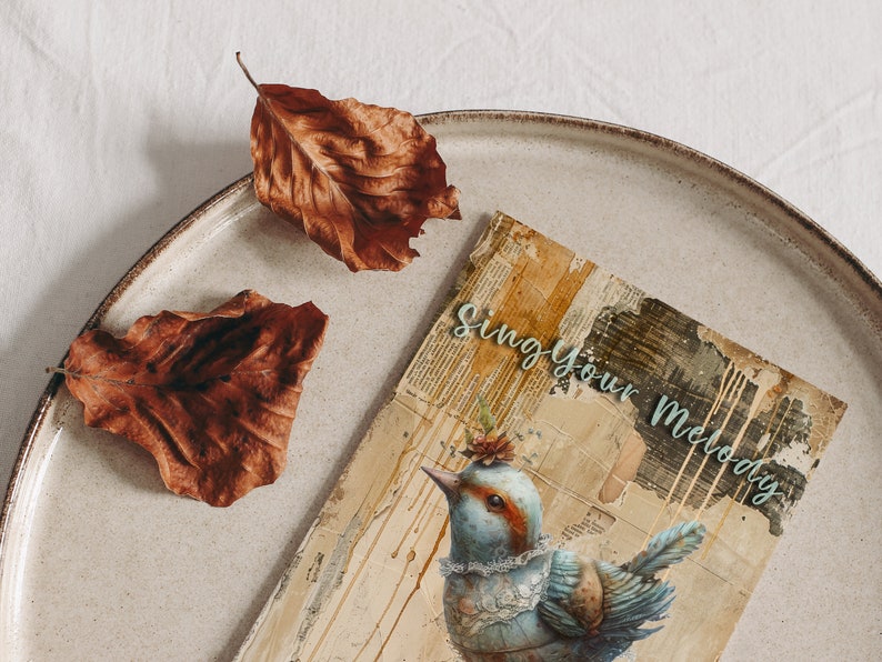 Puede incluir: Una tarjeta de estilo vintage con una ilustraci&oacute;n de un p&aacute;jaro azul y verde y el texto "Sing Your Melody". La tarjeta est&aacute; sobre un plato beige con dos hojas marrones.