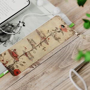 May include: A vintage-style bookmark with a whimsical illustration of teacups and saucers hanging from a tree branch. The bookmark is decorated with red flowers and has a beige tassel.