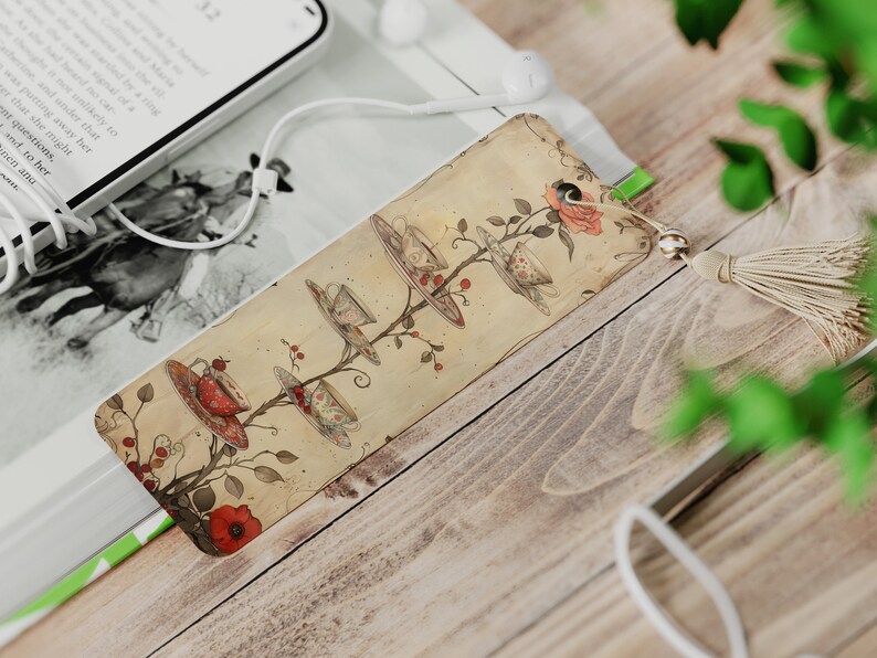 May include: A vintage-style bookmark with a whimsical illustration of teacups and saucers hanging from a tree branch. The bookmark is decorated with red flowers and has a beige tassel.