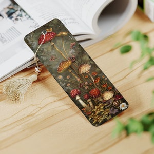 May include: A bookmark featuring a detailed illustration of red and white mushrooms with yellow stems growing in a forest setting. The bookmark is made of a dark brown material and has a tassel.