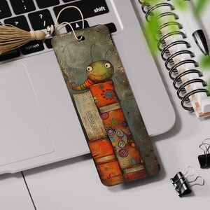 May include: A colorful bookmark featuring a cartoon bug with a big smile, standing on top of a stack of books. The bug is green with orange stripes and has large, round eyes. The books are orange, red, and brown with colorful designs.