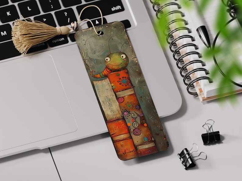 May include: A colorful bookmark featuring a cartoon bug with a big smile, standing on top of a stack of books. The bug is green with orange stripes and has large, round eyes. The books are orange, red, and brown with colorful designs.