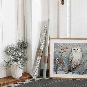 May include: A framed print of a snowy owl perched on a branch with red berries and white flowers. The owl has white feathers with black markings and yellow eyes. The background is a light blue watercolor wash.