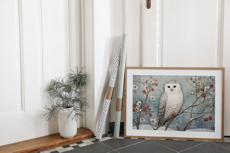 May include: A framed print of a snowy owl perched on a branch with red berries and white flowers. The owl has white feathers with black markings and yellow eyes. The background is a light blue watercolor wash.