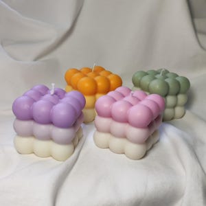 Op de afbeelding: Vier vierkante bellenkaarsen in verschillende kleuren: lavendel, oranje, roze en groen. Elke kaars heeft een witte basis en een bovenlaag van gekleurde bollen. De kaarsen zijn gerangschikt op een witte stoffen ondergrond.
