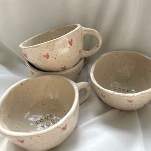 Op de afbeelding: Vier keramische mokken met gespikkelde glazuur en roze hartjes. Eén mok zegt "LOVE YOU" en een andere zegt "LOVE".