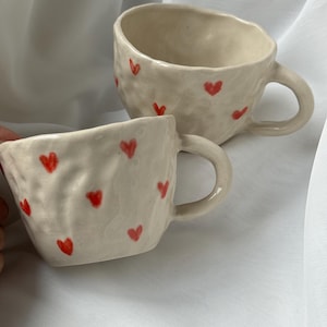 Op de afbeelding: Twee witte keramische mokken met rode hartjes. De mokken hebben een getextureerd, onregelmatig oppervlak.