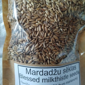 May include: A transparent bag filled with brown milk thistle seeds. The bag features a white label with the text "Mardadžu sēklas Blessed milkthistle seeds (Silybum marianum)" and a beaver illustration. The bag is set against a brown paper backdrop.