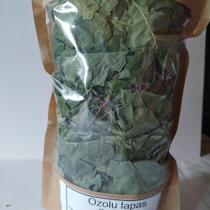 May include: A clear plastic bag filled with dried green oak leaves. The bag is sealed and contained within a brown paper bag. A white label on the front reads "Ozolu lapas Oak leaf" with a small illustration of a beaver.