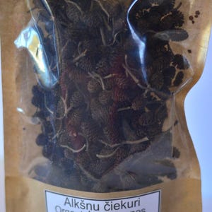 Puede incluir: Una bolsa de papel marrón con una ventana de plástico transparente que contiene conos de aliso secos. La bolsa tiene una etiqueta blanca que dice "Alksnu ciekkuri Organic alder cones 70g".