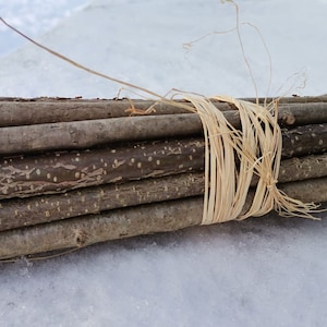Puede incluir: Un haz de pequeños palos de madera natural atados con rafia. Los palos son de varios tonos de marrón y gris, con textura de corteza visible. La rafia es de color beige claro. El paquete descansa sobre una superficie nevada.