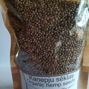 May include: A clear plastic pouch filled with small, round, brown and green hemp seeds. The label on the front reads: Kanepju sēklas, Organic hemp seeds, (Cannabis sativa), 200g. The pouch has a tan paper-like backing and a transparent front.