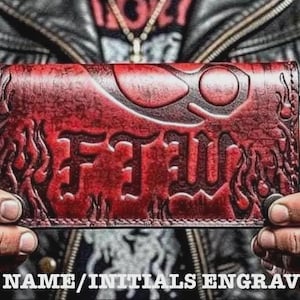 Op de afbeelding: Een rode leren portemonnee met een reliëf ontwerp en de letters "FEW". De portemonnee wordt voor een leren jas gehouden. De tekst "FREE NAME/INITIALS ENGRAVING" staat onderaan.