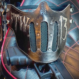 Può includere: Una maschera facciale in pelle marrone con la scritta "FTW" e il numero "13" in bianco. La maschera ha un design a rete sulla zona della bocca ed è rivettata. Una pistola nera è visibile sotto.