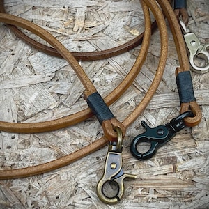 May include: Close-up of a tan leather dog leash with black and brass-colored metal hardware. The leash is coiled and has a simple design. The background is a light brown, textured surface.