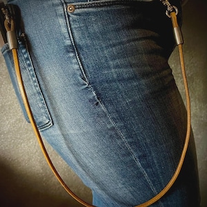 May include: A light brown leather wallet chain with silver-toned metal accents. The chain is attached to a blue denim jean pocket and loops down. The leather is a warm, natural color.