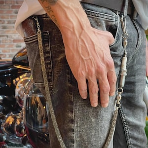 Könnte beinhalten: Eine silberne Metall-Geldbörsenkette aus einer Fahrradkette und einer Zündkerze. Die Kette ist an einer grauen Jeans befestigt. Die Person trägt einen schwarzen Gürtel und ein weißes T-Shirt.