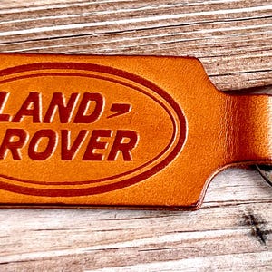 May include: A tan leather keyring featuring a rectangular shape with rounded corners and a metal split ring. The surface is embossed with an oval border and the text LAND ROVER in a bold, dark brown font centred within the oval design.