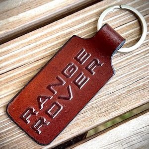 May include: A rectangular brown leather keychain featuring the embossed text RANGE ROVER in a bold, sans-serif font. The leather tag is attached to a circular silver-toned metal split ring, showcasing a smooth, polished finish on the leather surface.