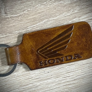 Op de afbeelding: Een rechthoekige bruine leren sleutelhanger met het in reliëf aangebrachte Honda-vleugellogo en het woord HONDA in hoofdletters. De sleutelhanger bevat een ronde metalen ring aan één uiteinde en heeft een gladde, gepolijste lederen textuur.