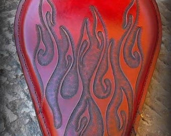 May include: A red leather motorcycle seat with a flame design. The seat has a teardrop shape and features a carved flame pattern in shades of red, orange, and purple. The edges are stitched with a contrasting thread.