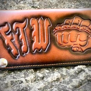 Könnte beinhalten: Ein rechteckiges Portemonnaie aus braunem Leder mit dunklem Farbverlauf und sichtbaren Nähten an den Rändern. Die Vorderseite zeigt die geprägten Buchstaben FTW sowie eine Grafik einer geballten Faust mit Schlagring, befestigt mit zwei silbernen Druckknöpfen.
