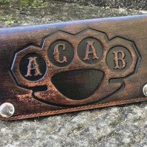 Op de afbeelding: Bruin leren portemonnee met een boksbeugelontwerp. Het ontwerp bevat de letters "A", "C", "A" en "B" in afzonderlijke cirkels. De portemonnee heeft een drukknoopsluiting en is gestikt met oranje draad.