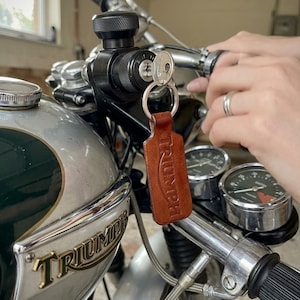 以下が含まれることがあります： 茶色の革製 Triumph キーホルダーがオートバイのキーにぶら下がっています。キーホルダーは長方形で、Triumph のロゴがエンボス加工されています。オートバイはシルバーの燃料タンク、クロムハンドルバー、黒いゲージを備えています。