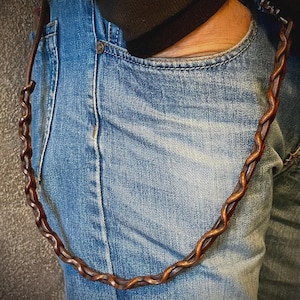 May include: A brown leather wallet chain with a twisted rope design hangs from a blue denim jean pocket. The chain is a rich, dark brown color and appears to be made of high-quality leather. The jeans are a light blue wash.