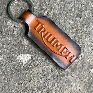 Può includere: Un portachiavi Triumph in pelle con un anello argentato. Il portachiavi rettangolare con bordi arrotondati presenta il logo Triumph in rilievo arancione. La pelle ha un effetto sfumato, che passa dal marrone al nero.