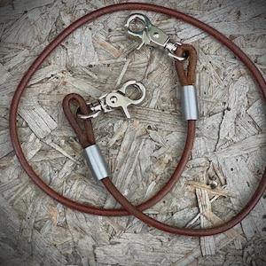 May include: A brown leather leash with two silver-colored metal clasps. The leash is circular and appears to be made of a single piece of leather. The clasps are attached to the leather with a small loop and a silver metal tube.