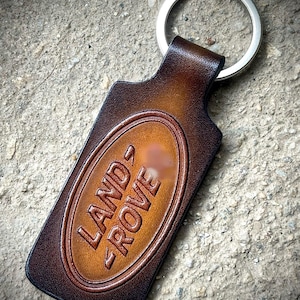 Pode incluir: Um porta-chaves de couro castanho com o texto em relevo "LAND ROVE" num oval castanho claro. O porta-chaves tem um anel de metal prateado preso no topo. O couro tem um aspeto texturizado.