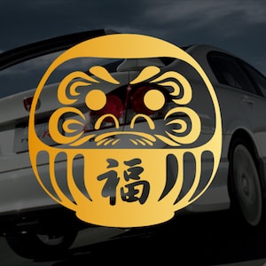 May include: A gold daruma doll sticker with black details and the Japanese character for "fortune" on a white car.