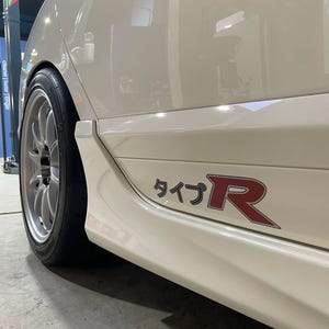 May include: A white car side panel featuring a Type R decal with Japanese characters and a large red letter R. The vehicle has a silver multi-spoke wheel with a black tire, and the bodywork shows a smooth, glossy finish with sharp side skirt lines.