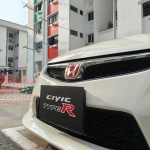 May include: A white Honda Civic Type R car with a black front grill and a red Honda emblem. A black license plate with the text "CIVIC TYPE R" in white and red is visible.