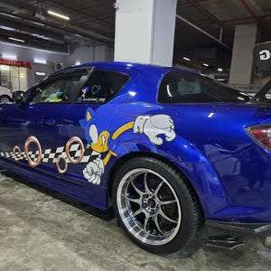 Peut inclure: Une voiture de sport bleu vif arborant un grand graphique du personnage Sonic le hérisson sur le côté, accompagné d'anneaux dorés et d'un motif à damier noir et blanc. Le véhicule est équipé d'un aileron arrière proéminent et de jantes argentées.
