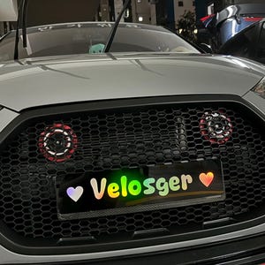 Puede incluir: Primer plano de la parrilla delantera negra en forma de panal de un coche plateado. La matrícula muestra "Velosger" en colores arcoíris, con un corazón morado y un corazón rojo. Luces circulares rojas y negras a cada lado de la parrilla.