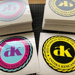 May include: Stacks of white stickers with a black and yellow circle design. The circle has the text "SEJUTA IMPIAN MOGA PUTIK MENJADI ANDIKA KENCANA" and the letters "ak" in yellow.
