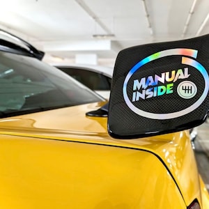 Pode incluir: Um carro amarelo com um spoiler preto que tem um adesivo que diz "MANUAL INSIDE" com um símbolo de câmbio de marchas.