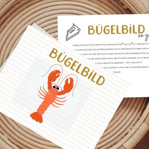 Könnte beinhalten: Eine weiße Karte mit einem Cartoon-Hummer-Design und dem Wort "BÜGELBILD" in goldfarbenen Buchstaben. Eine zweite Karte enthält Anweisungen zum Aufbringen des Designs mit einem Bügeleisen. Der Hummer ist orange mit schwarzen Augen.