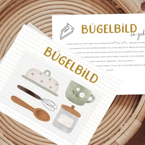 Könnte beinhalten: Eine weiße Karte mit dem Text "Bügelbild so geht's" in goldenen Buchstaben. Die Karte enthält Anweisungen zum Aufbügeln eines Transfers. Die Karte liegt auf einem geflochtenen Korb mit einer weiß-braunen Karte mit verschiedenen Küchenutensilien.