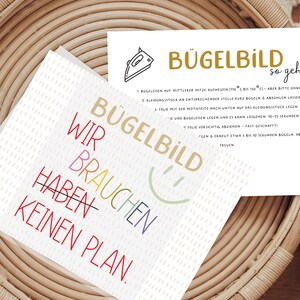 Könnte beinhalten: Zwei weiße Karten mit Text und einem Smiley-Gesicht werden auf einem Weidenkorb präsentiert. Eine Karte zeigt "Bügelbild" in Gold und "Wir brauchen keinen Plan" in farbigem Text. Die andere Karte enthält Anweisungen und die Worte "Bügelbild so geht's".