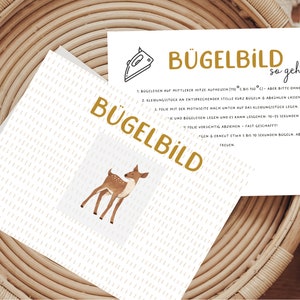 Könnte beinhalten: Eine weiße Karte mit einem Hirsch-Motiv und dem Wort "BÜGELBILD" in goldfarbenen Buchstaben. Eine zweite Karte mit Anweisungen und den Worten "BÜGELBILD so geht's" ist ebenfalls sichtbar. Die Karten liegen auf einer gewebten Oberfläche.
