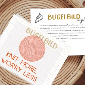 Könnte beinhalten: Eine weiße Karte mit dem Text "Knit More, Worry Less." in Orange und Rot. Eine rote und weiße Wollkugel befindet sich in der oberen linken Ecke. Die Karte enthält auch den Text "Bügelbild" in Gold und Anweisungen zum Aufbügeln des Designs.